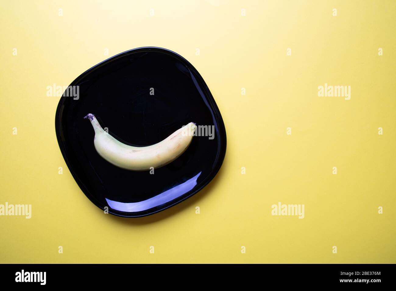Ein modernes pastellfarbenes Bild einer Banane und ein dekorativer schwarzer quadratischer Teller mit pastellgelbem Hintergrund, flacher Lage, Draufsicht Stockfoto