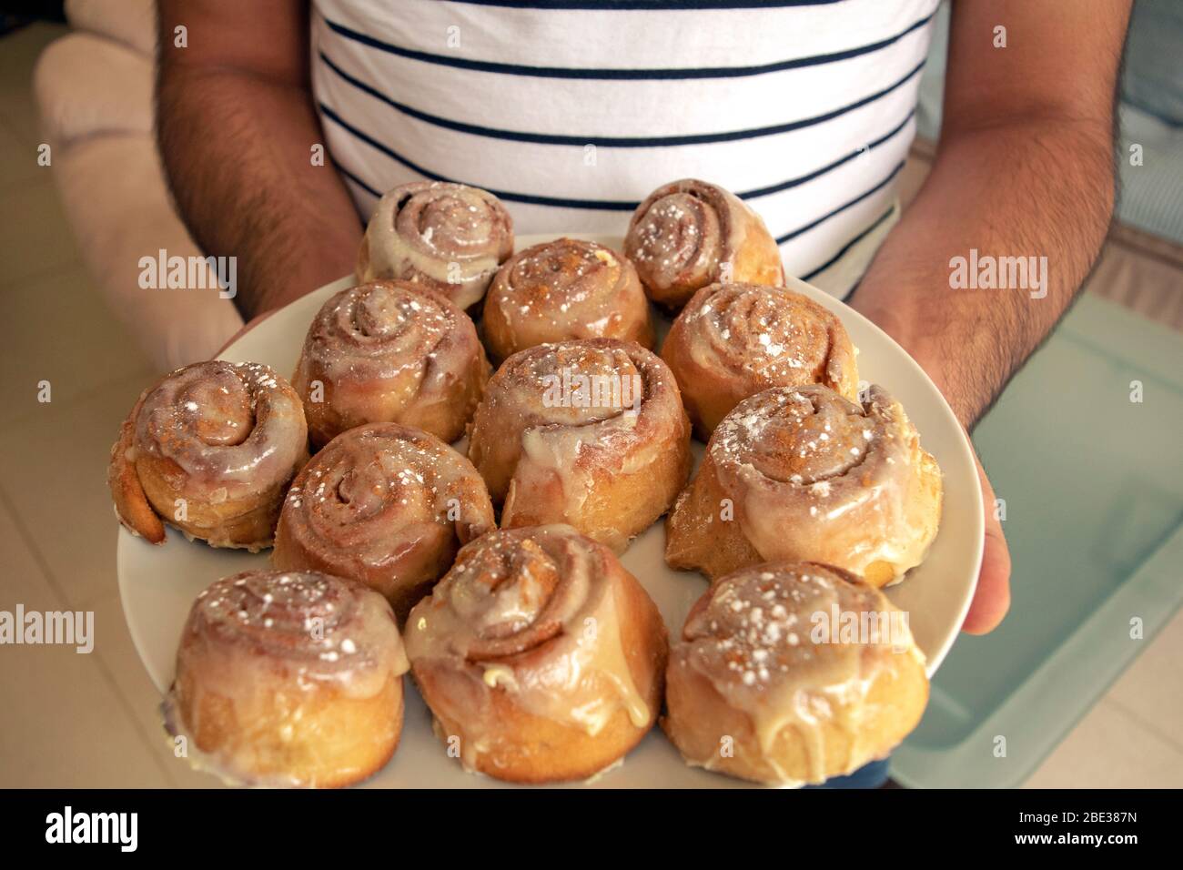 Ein Mann hält hausgemachte frisch gebackene Cinnabon Buns mit Sahne-Sauce und Zimtpulver auf einem großen weißen Teller auf hellem unscharfen Hintergrund. Hausgemachte Bäckerei. Stockfoto