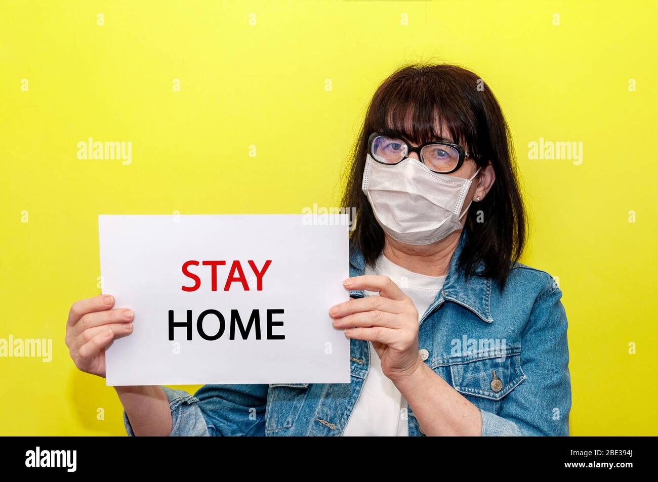 COVID-19. Ältere Frau in schützender Gesichtsmaske hält Schild zu Hause bleiben auf gelbem Hintergrund. Anti-Coronavirus-Konzept. Kopieren Sie Den Bereich Für Ihren Text. Stockfoto