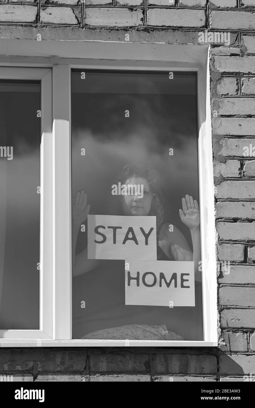 Eine Frau sitzt neben einem Fenster mit der Inschrift zu Hause bleiben, Haus Quarantäne Konzept während einer Coronavirus-Pandemie, schwarz-weiß-Foto. Stockfoto