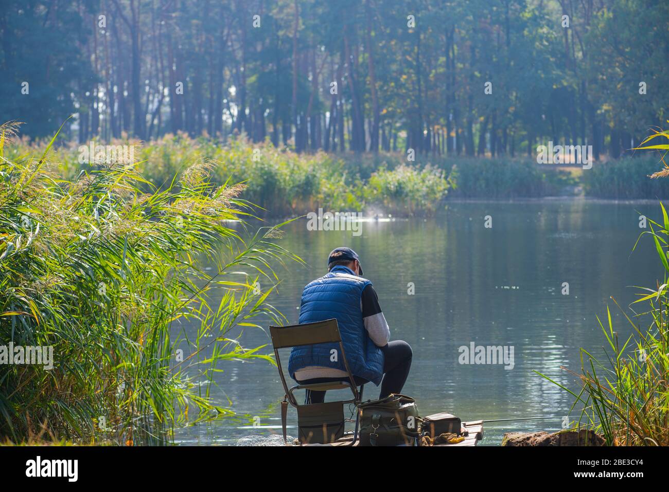 Ein Fischer mit einer Rute, der in den Strahlen der Herbstsonne am ruhigen Ufer eines Waldsees sitzt. Angelzubehör. Das Konzept der Outdoor-Aktivität Stockfoto