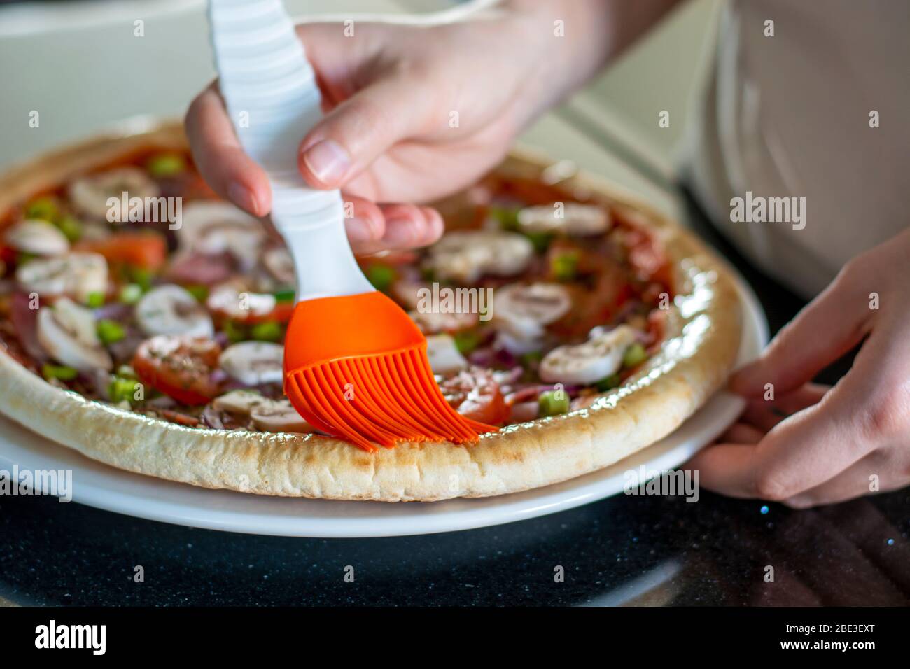 Hausgemachte Pizza mit Gemüse. Der Prozess der Herstellung von Pizza zu Hause. Hände von Frau, die Pizza macht und die Ränder der Kruste mit Olivenöl putzt. Stockfoto