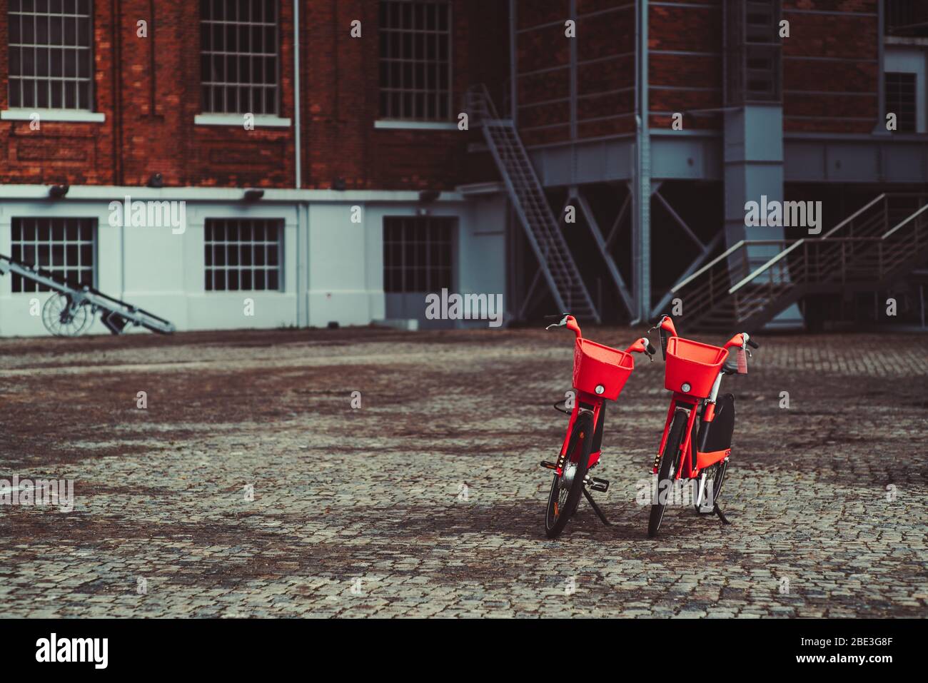 Zwei leuchtend rote Leihfahrräder, die auf einem Pflasterstein vor einem Backsteingebäude geparkt sind; zwei Gemeinschaftsfahrräder mit Metallkörben am Steuerstand im Freien Stockfoto