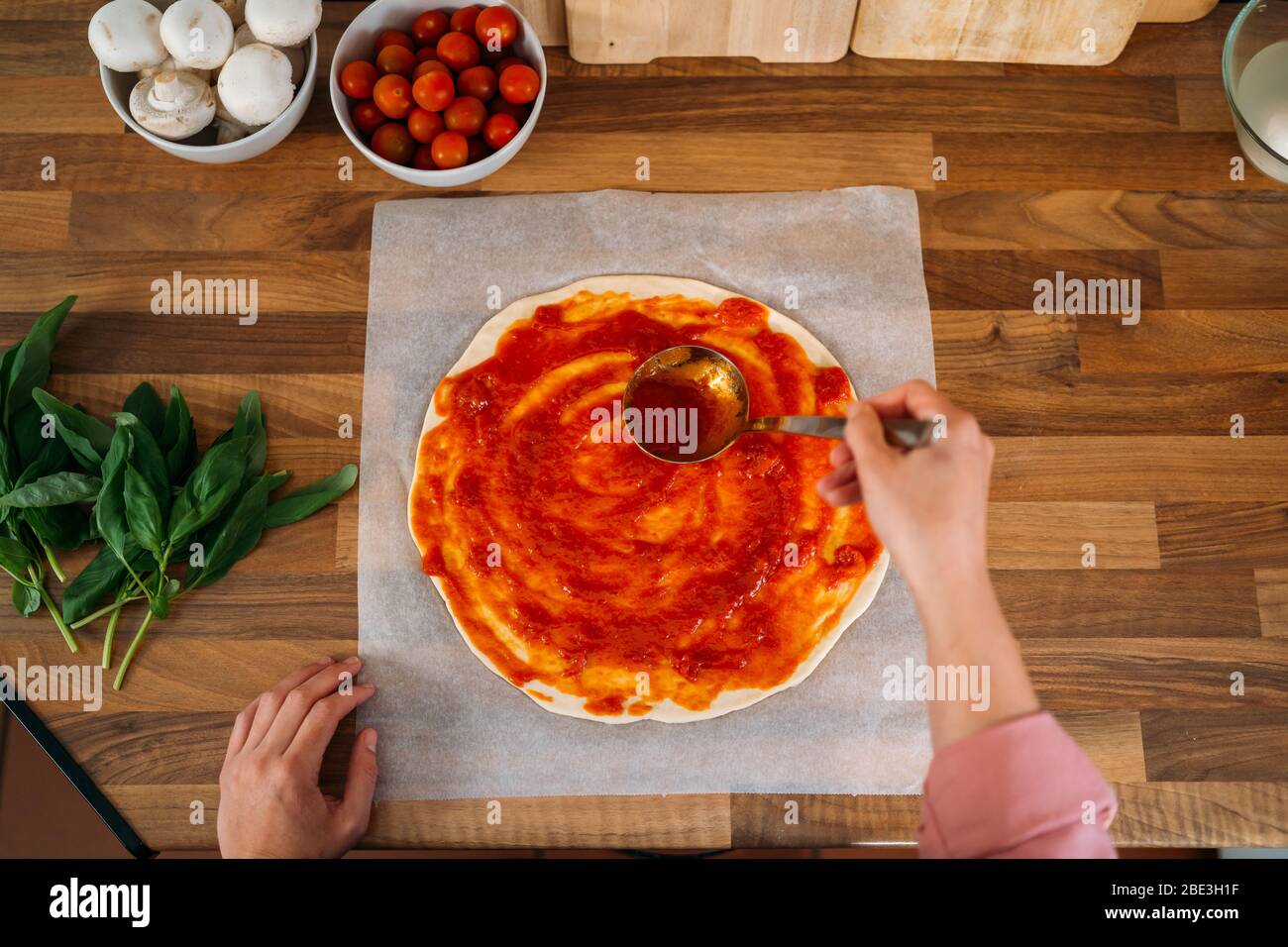 Draufsicht auf die Hände einer Frau, die Tomatensauce zu einem Pizzateig auf einer Holzarbeitspfläche hinzufügt. Roher Teig für die Pizzazubereitung mit Zutat: Tomatensauce, m Stockfoto