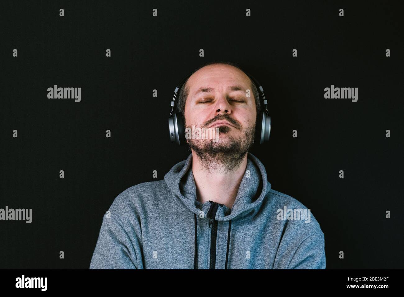 Bärtiger entspannter Mann im Hoodie, der mit Kopfhörern vor schwarzem Hintergrund steht, Musik hört, Kopf und Schultern Stockfoto