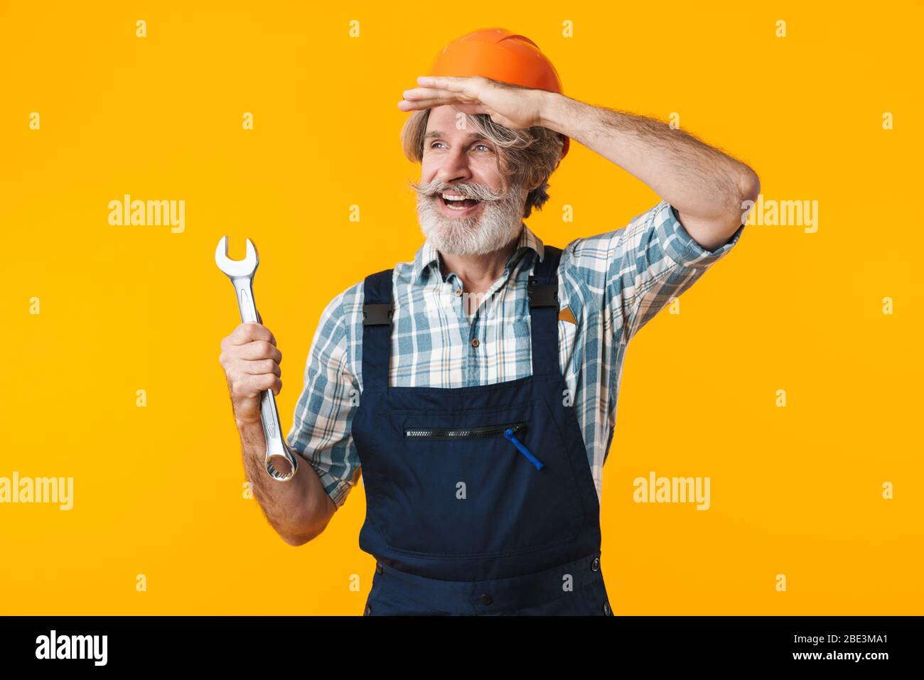 Bild von positiven optimistischen älteren grauhaarigen bärtigen Mann Builder in Helm posiert isoliert über gelbe Wand Hintergrund halten Schraubenschlüssel suchen asid Stockfoto