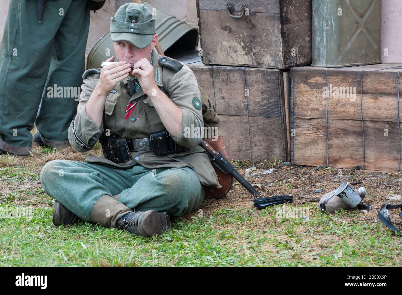 Ein Soldat, der auf dem Boden sitzt, eine Mundharmonika spielt, in einer grünen Uniform gekleidet bei einer Nachstellung des Zweiten Weltkriegs. Stockfoto