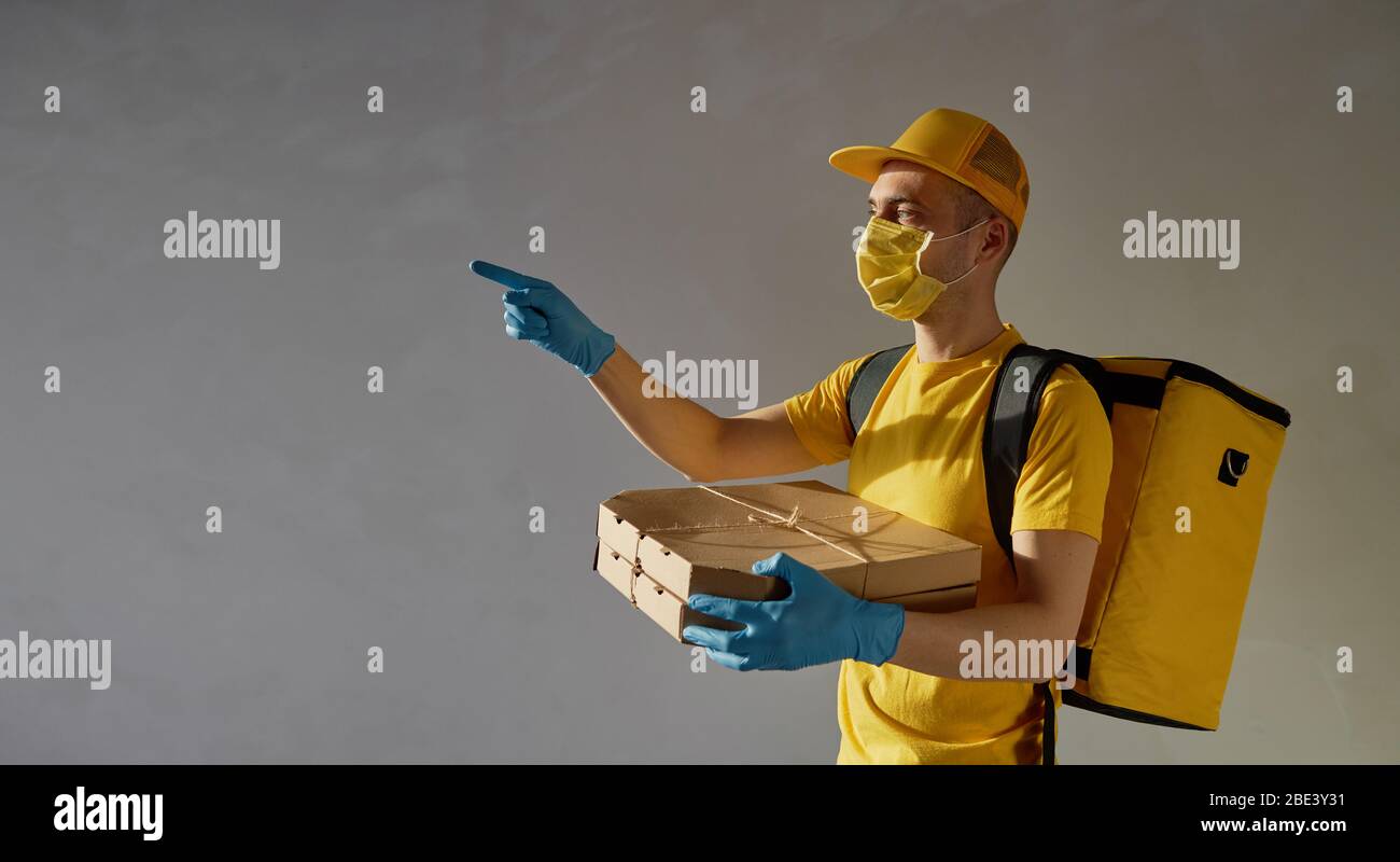 Sichere Lieferung von Lebensmitteln. Kurier in gelber Uniform, Schutzmaske und Handschuhe liefert während der Coronovirus-Quarantäne Essen zum Mitnehmen. Nach links zeigend. Stockfoto