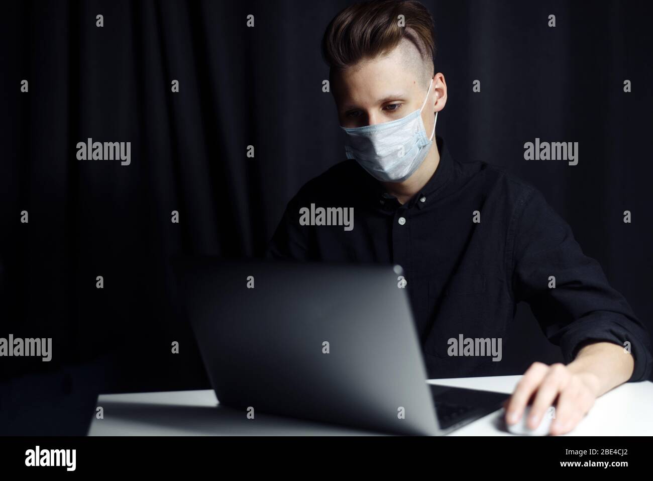Ein junger Freiberufler arbeitet von zu Hause aus. Der Typ sitzt zu Hause in einer Schutzmaske, selbstisolierend und sitzt in einem Laptop. Stockfoto