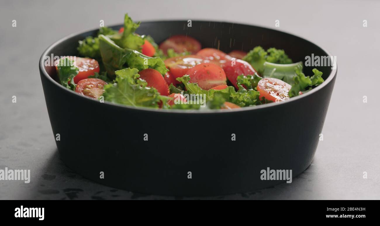 Man würzt Salat mit Tomaten und Grünkohl in schwarzer Schüssel Auf Betonoberfläche Stockfoto