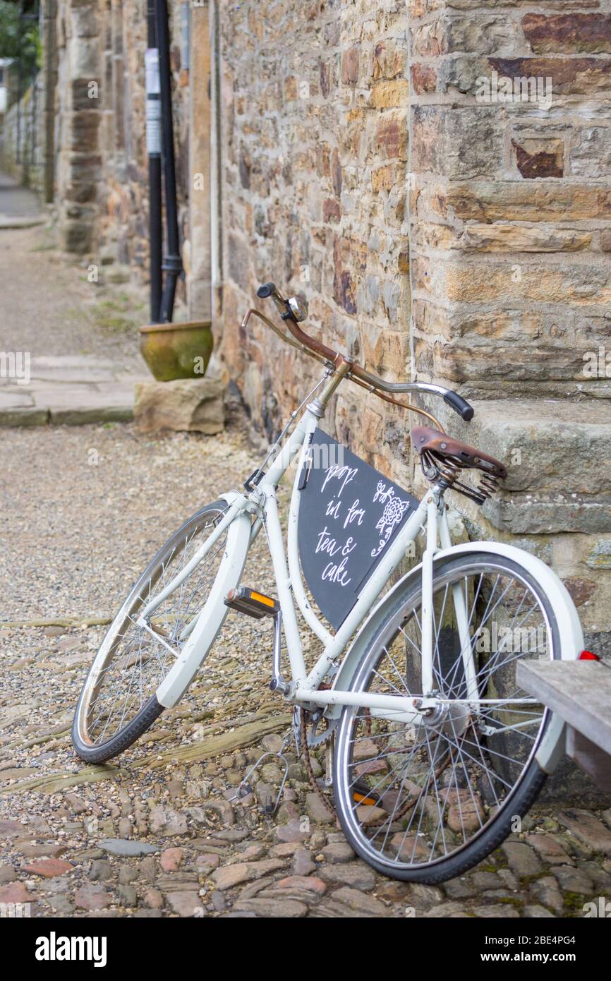 Altmodisches, weiß gestrichenes Damenfahrrad, das an einer hellgelben Steinaußenwand vor einem Café in einem hübschen Dorf befestigt war Stockfoto