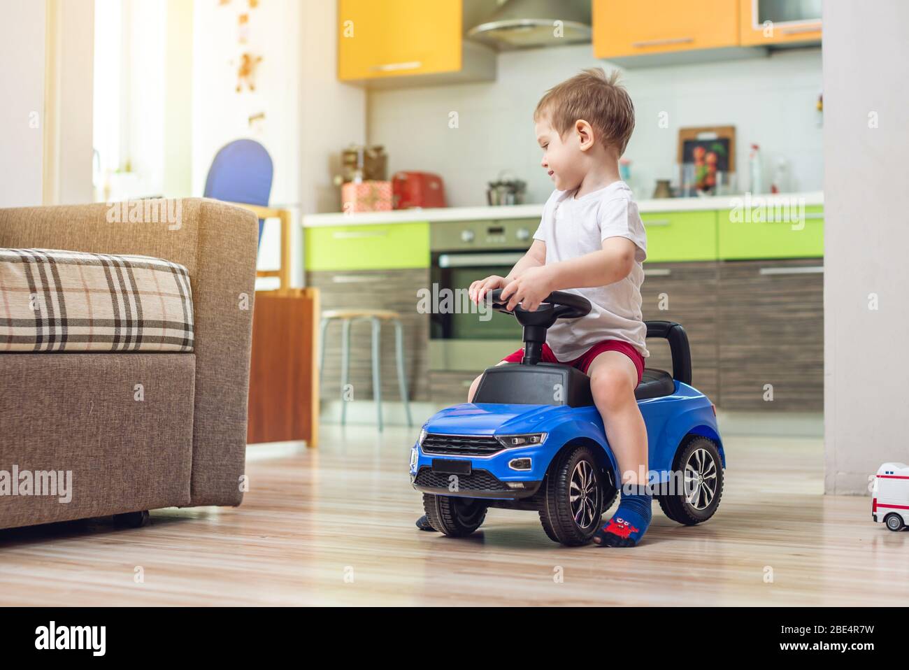 Glücklicher kleiner Junge fährt auf einem Spielzeugauto in der Wohnung. Aktivität für ein Kind, wenn zu Hause. Organisation der Unterhaltung für das Kind. Stockfoto