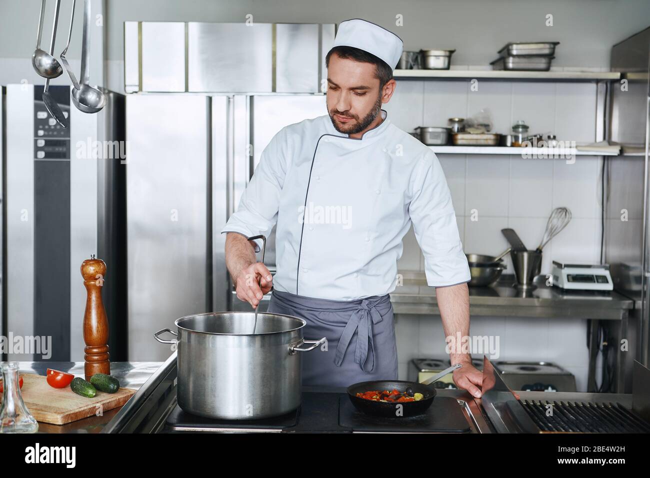 Konzentrierter kaukasischer Koch in Uniform macht seinen Job. Er kocht die notwendigen Zutaten für sein Signature-Gericht. Vorderansicht. Kochkonzept Stockfoto