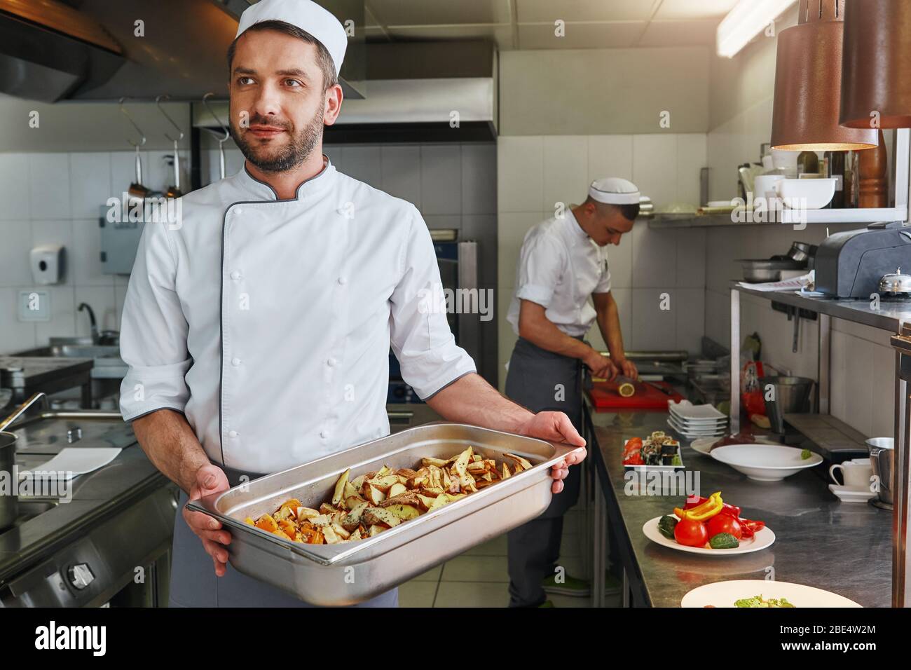 Lächelnder kaukasischer Koch in Uniform macht seinen Job. Er bereitet die notwendigen Zutaten für sein Signature-Gericht zu. Vorderansicht. Kochkonzept Stockfoto