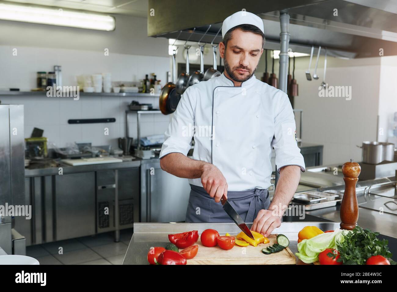 Konzentrierter kaukasischer Koch in Uniform macht seinen Job. Er zerkleinert Gemüse für sein charakteristisches Gericht. Kochkonzept Stockfoto