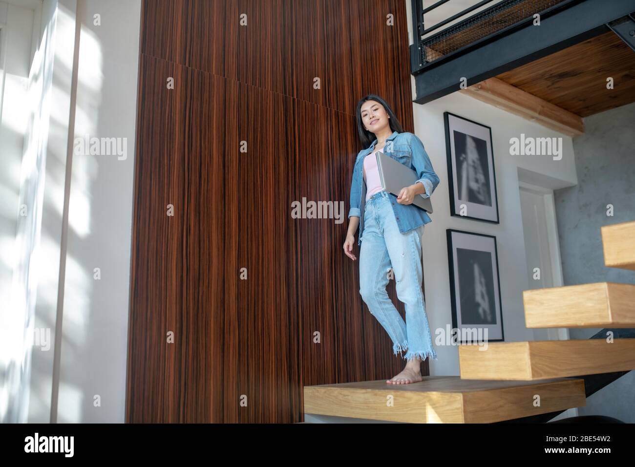 Junge Frau, die auf Treppen steht, Laptop unter dem Arm hält und sich an die Wand lehnt Stockfoto