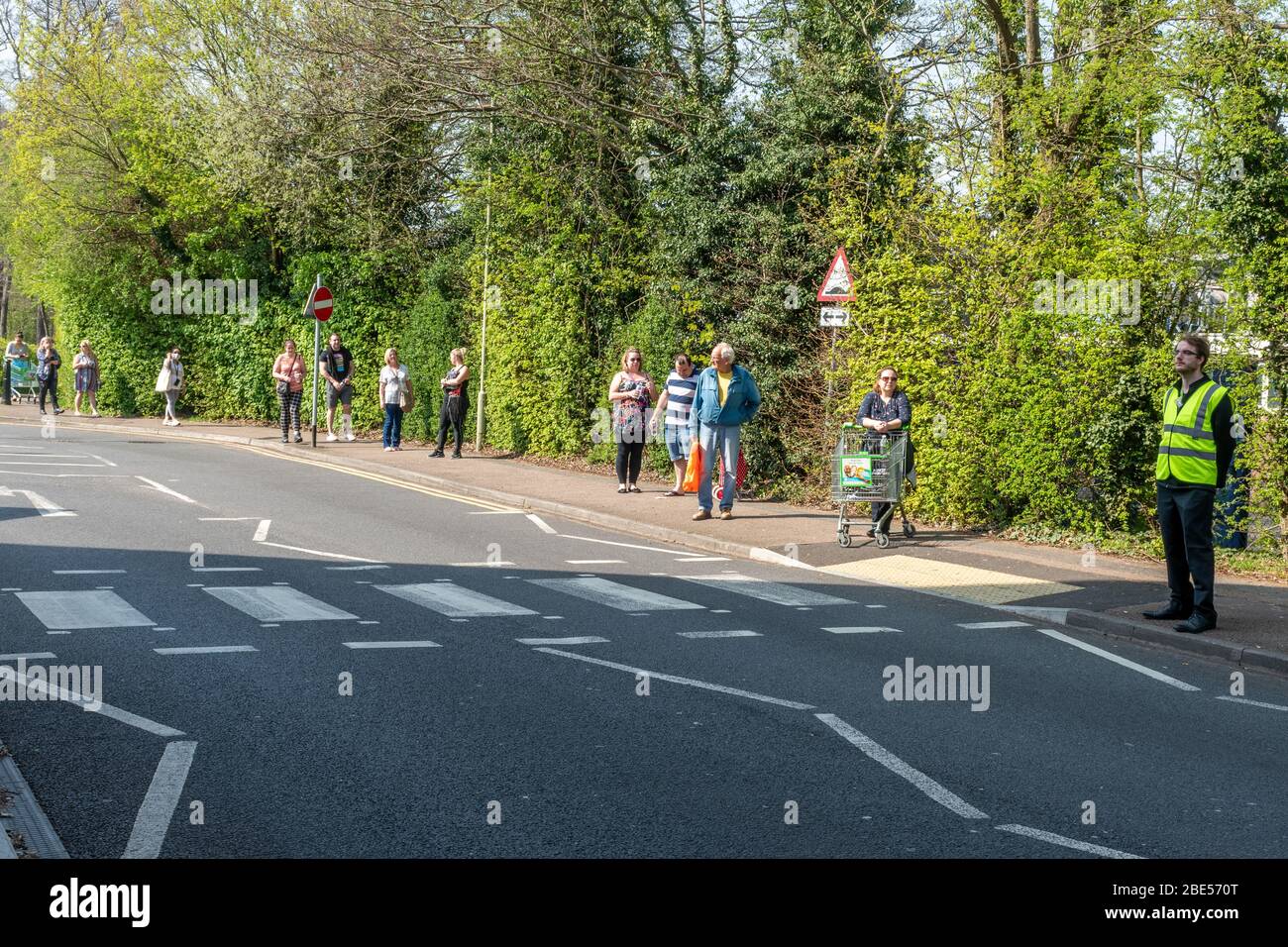 10. April 2020. Während der Coronavirus Covid-19-Pandemie von 2020 gibt es in Großbritannien eine Sperre, und die Menschen erlauben nur aus bestimmten Gründen, ausgehen zu dürfen. Einschließlich Lebensmitteleinkäufe. Eine lange Schlange, um in den Asda-Supermarkt in Farnborough, Hampshire, zu gehen, um nach lebensnotwendigen Vorräten zu kaufen. Menschen, die Schlange stehen, beobachten soziale Distanzierungen und stehen etwa zwei Meter voneinander entfernt. Ein Mitarbeiter hilft dabei, die Anzahl der Personen zu kontrollieren, die das Coffee House betreten, und zwar „One-in- und One-Out“-Basis. Stockfoto