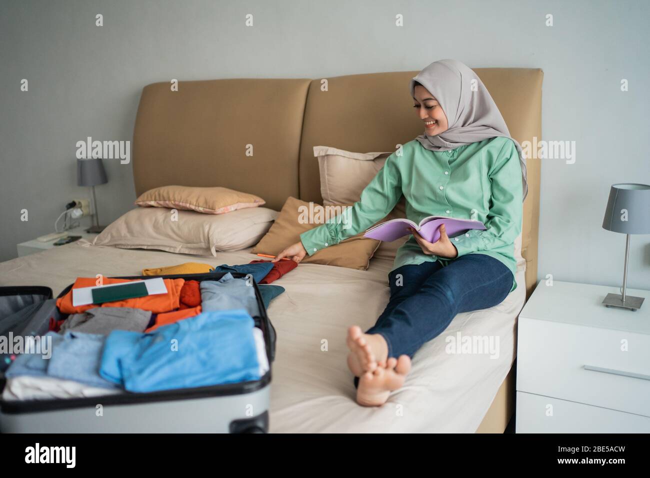 Verschleierte Frau, die auf dem Bett sitzt, während sie die Liste der Gegenstände betrachtet, die im Urlaub weggenommen werden sollen Stockfoto