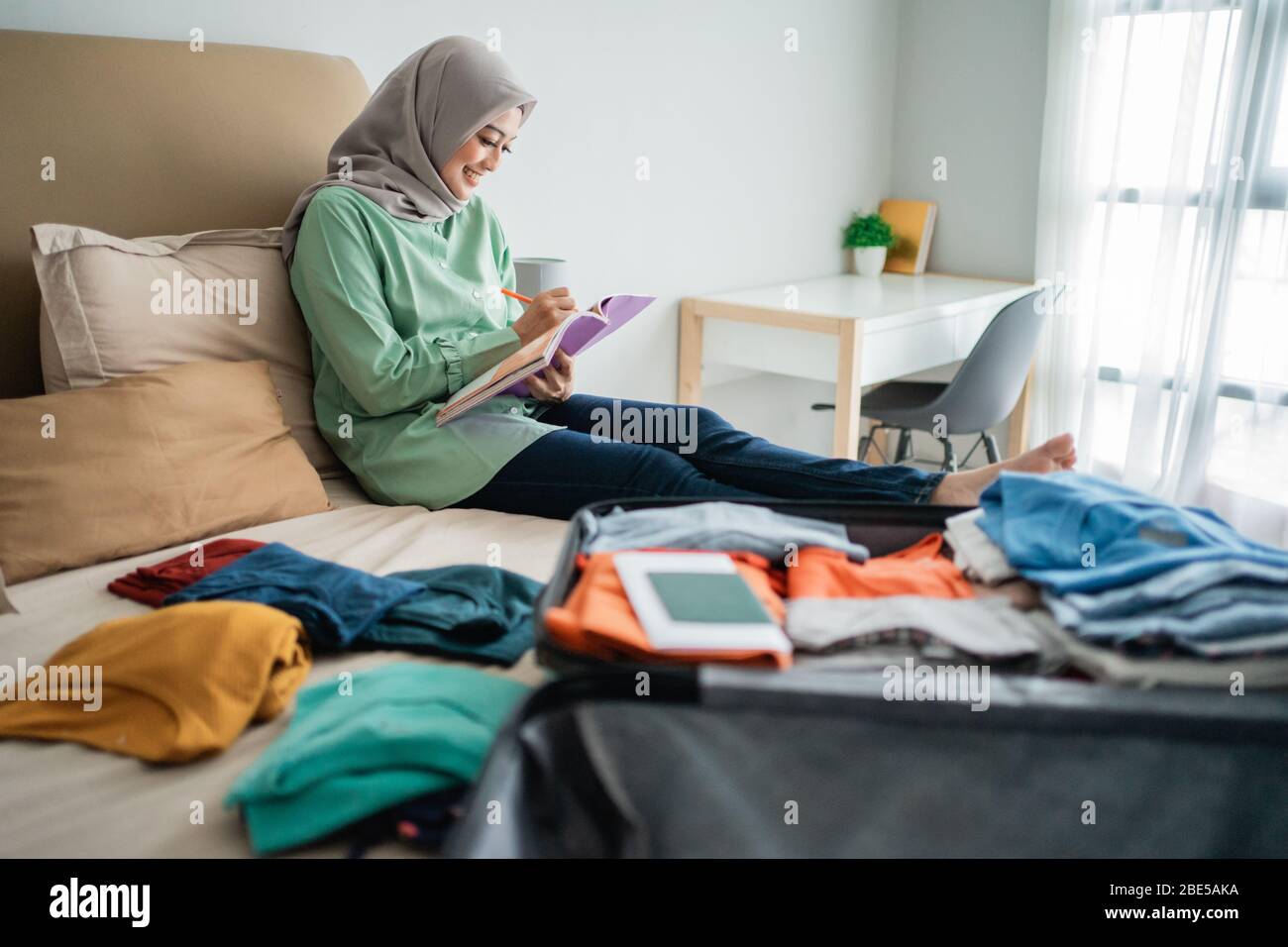 Verschleierte Frau, die auf dem Bett sitzt, während sie die Liste der Gegenstände betrachtet, die im Urlaub weggenommen werden sollen Stockfoto