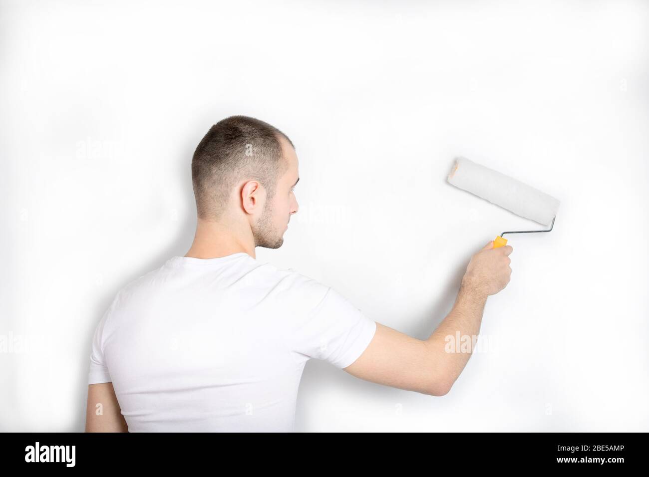 Ein Typ in einem weißen T-Shirt malt mit einer Farbwalze eine Wand. Stockfoto