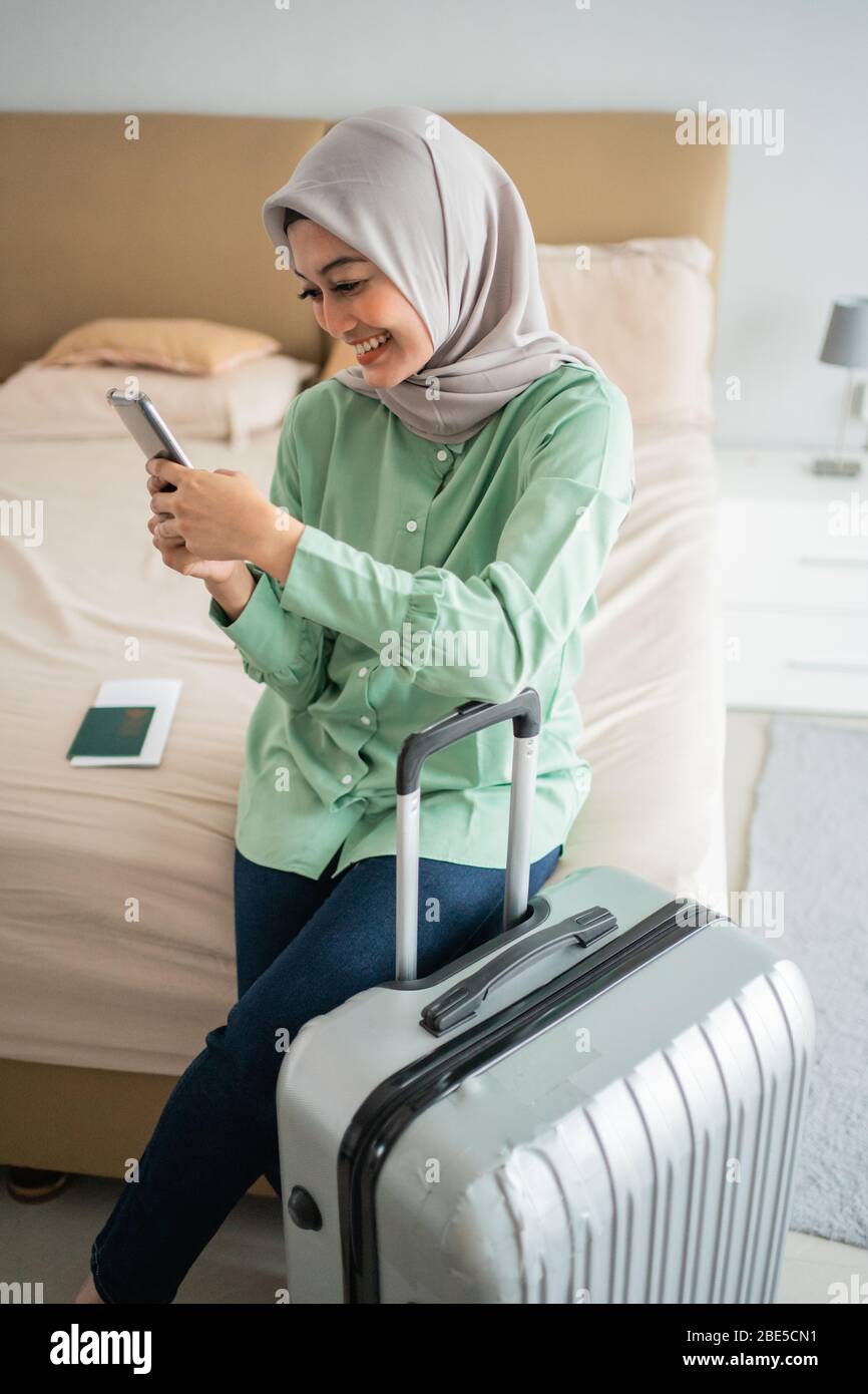 Die verschleierte Frau saß auf dem Bett und hielt ihr Smartphone und ihren Koffer, bevor sie in den Urlaub ging Stockfoto