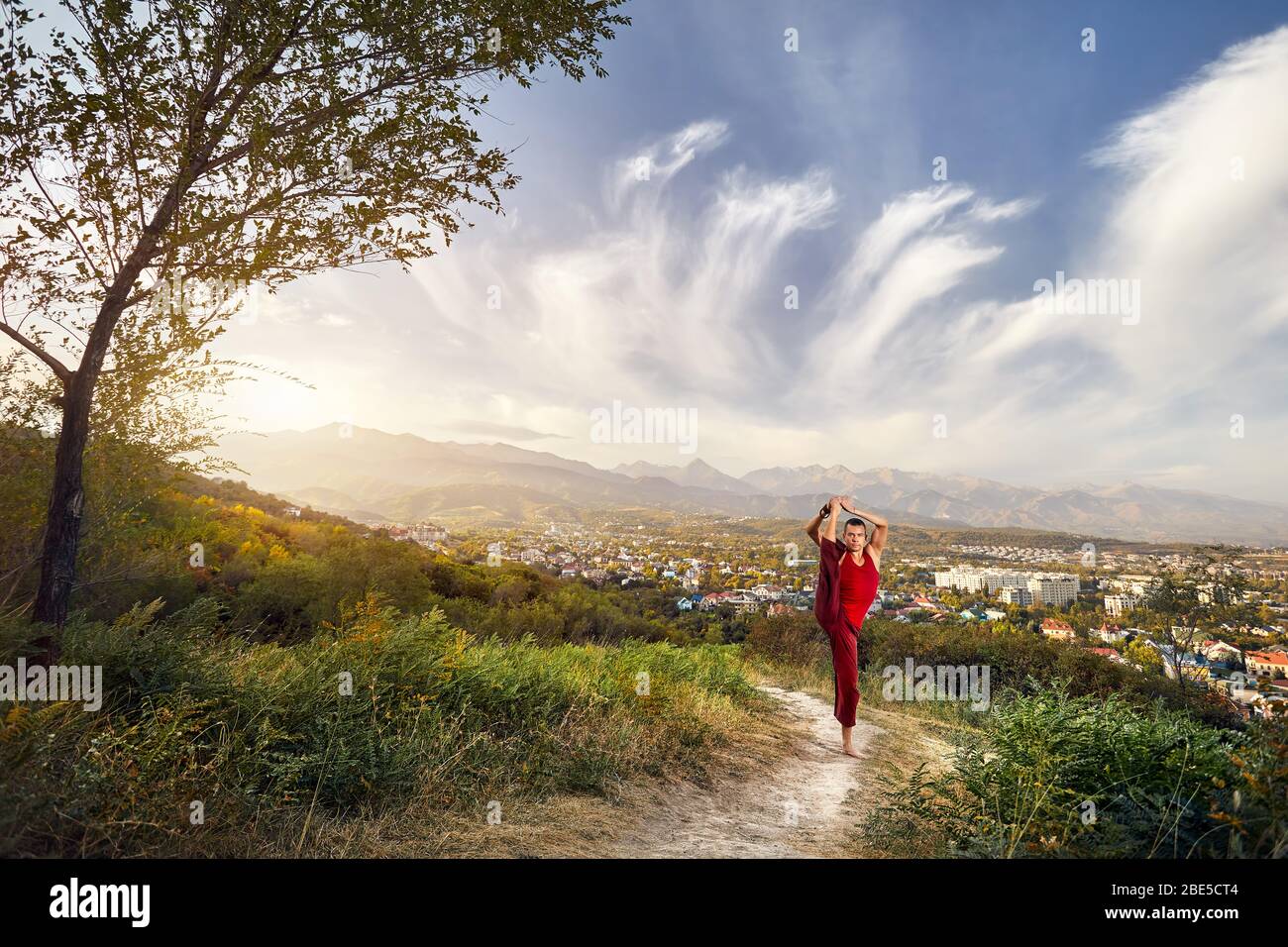 Passen Mann im roten Kostüm leg split Asana Yoga im Park mit die Stadt und die Berge im Hintergrund in Kasachstan Stockfoto