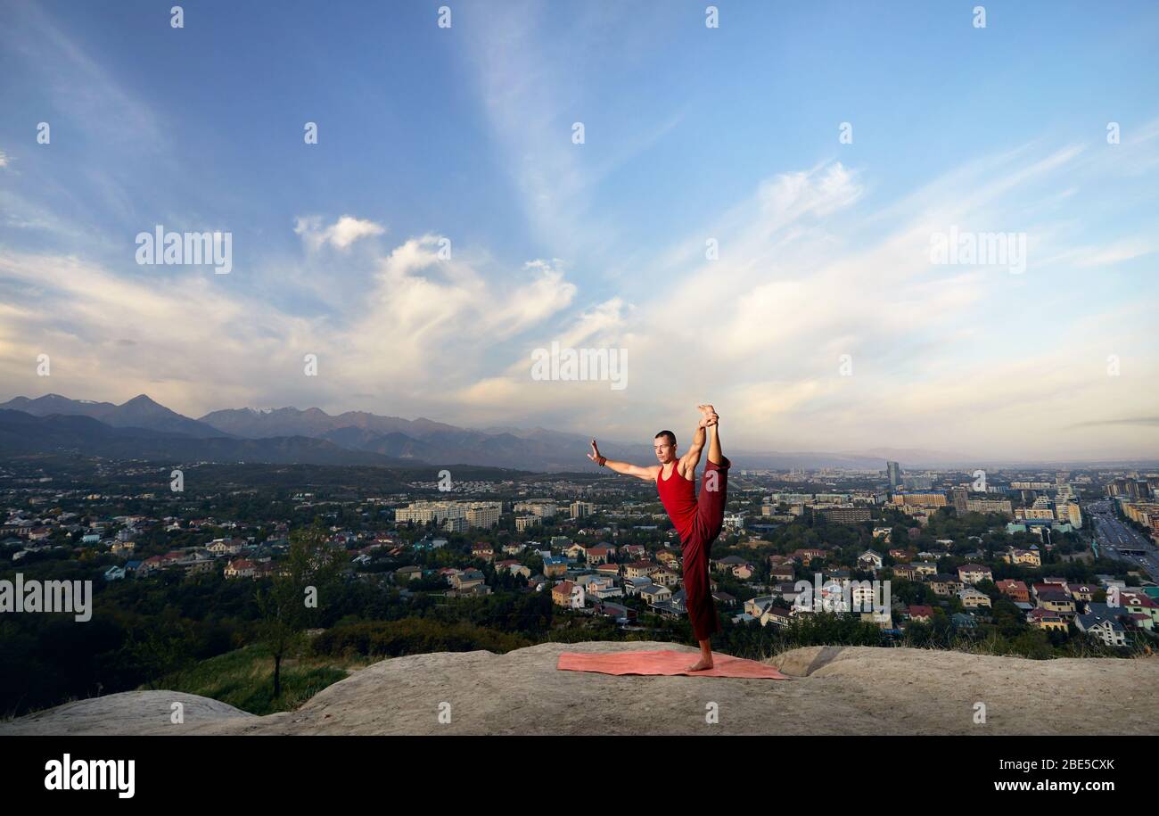 Passen Mann im roten Kostüm Yoga balance Asana im Park mit die Stadt und die Berge im Hintergrund in Almaty, Kasachstan Stockfoto
