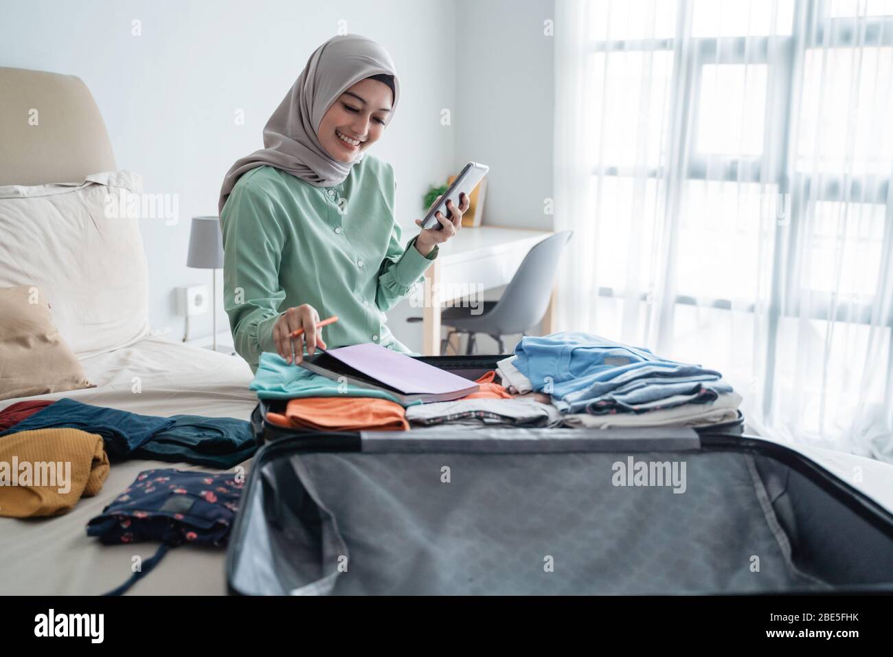 Hijab-Frau, die auf dem Bett sitzt, während sie die Liste der Gegenstände betrachtet, die im Urlaub weggenommen werden sollen Stockfoto