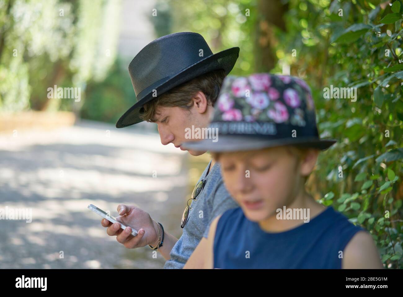 Zwei Teenager-Brüder in den Sommerferien, einer von ihnen hält ein Handy unter Bäumen im Grünen eines Parks sitzen Stockfoto