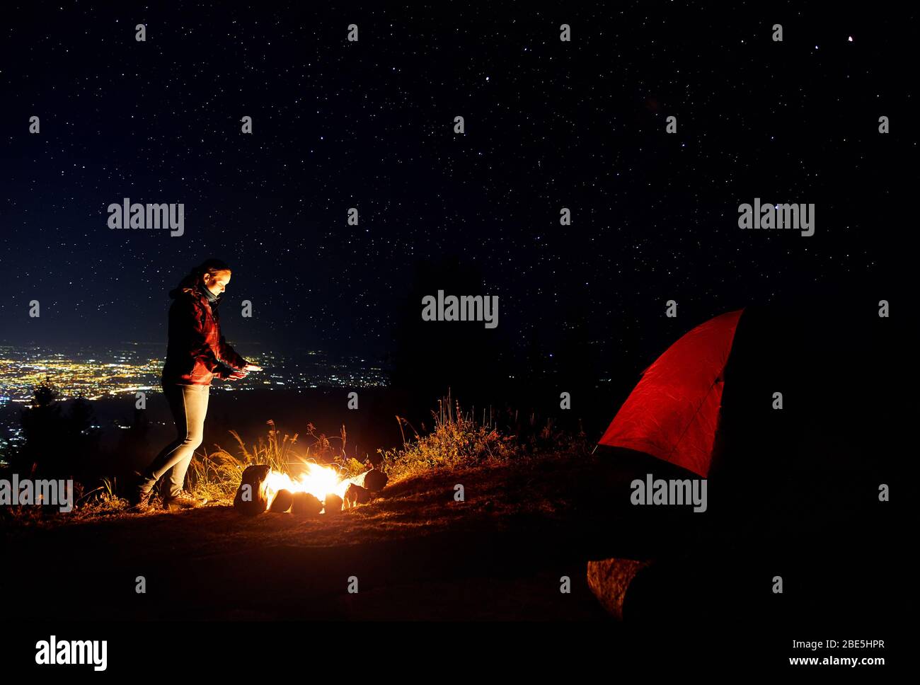 Schöne Wanderer Mädchen warme Hände in der Nähe von Lagerfeuer unter Sternenhimmel mit Sterne und die Lichter der Stadt im Hintergrund. Stockfoto