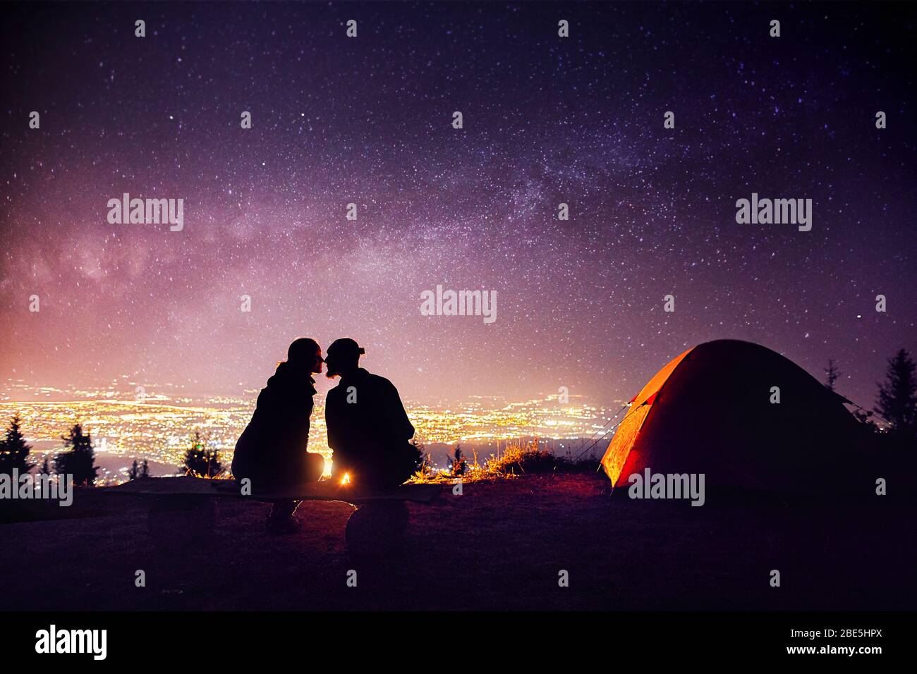 Glückliches Paar in silhouette Küssen in der Nähe von Lagerfeuer und orange Zelt. Nachthimmel mit milchstrasse Sterne und die Lichter der Stadt im Hintergrund. Stockfoto