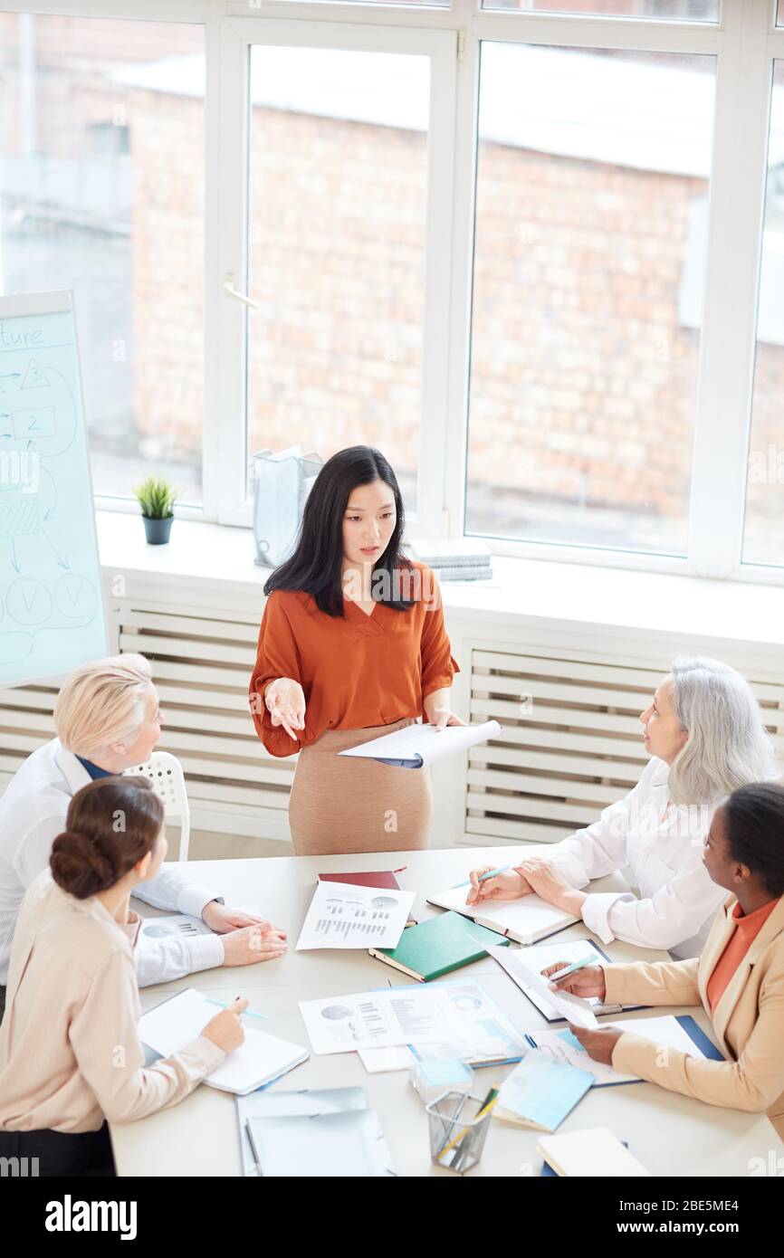 Vertikales Porträt einer erfolgreichen asiatischen Geschäftsfrau, die während eines Meetings in CO vor einer Gruppe von Kolleginnen am Tisch steht Stockfoto