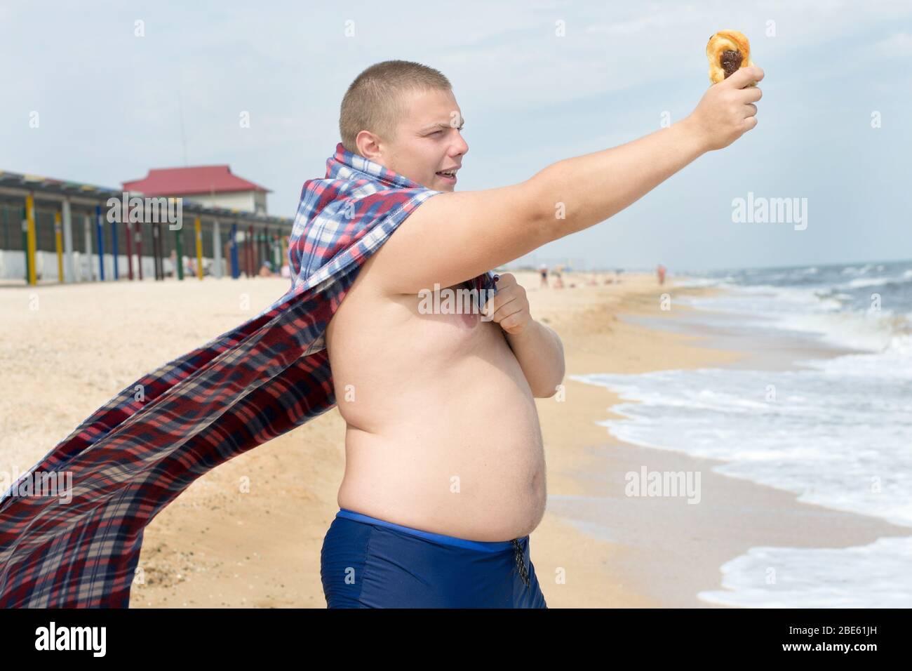 Am Strand steht ein witziger fetter Mann-Superheld mit Plaid statt Mantel und Bun in der Hand. Stockfoto