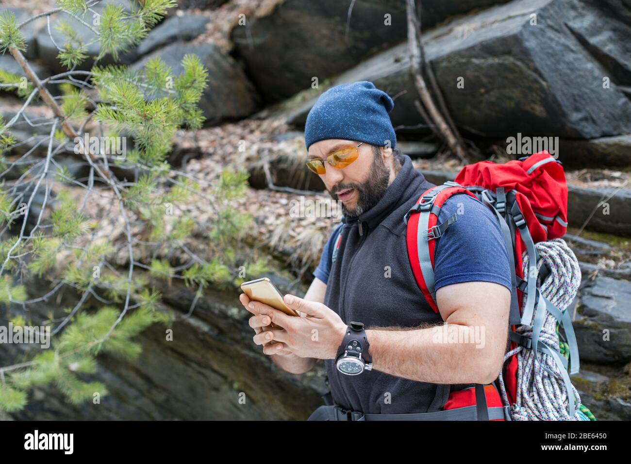 Ein Mann Tourist mit Rucksack steht in der Nähe eines bloßen Steinfelsen und Myotrite auf dem Bildschirm eines Mobiltelefons. Sucht mit dem GPS-Navigator nach einem Weg. Th Stockfoto