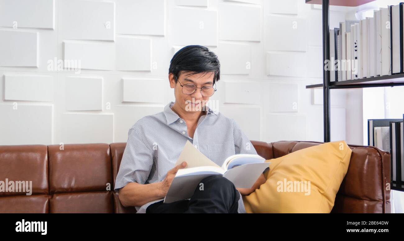Senior Asian Mann Lesen Buch auf Sofa im Wohnzimmer zu Hause, Porträt von asiatischen älteren Mann ist entspannend und Glück mit Lesen Sie ein Magazin zu Hause Stockfoto
