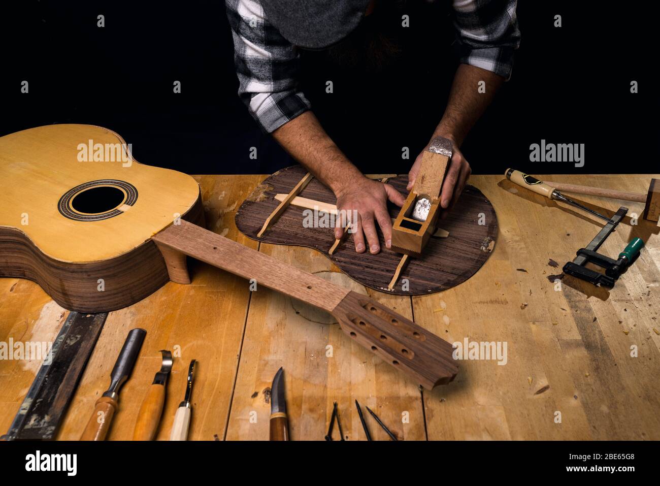 Mann trägt Mütze und kariertes Hemd, das Gitarre macht. Planen Gitarre zurück. Werkstatt von Luthier. Dunkelschwarzer Hintergrund. Stockfoto