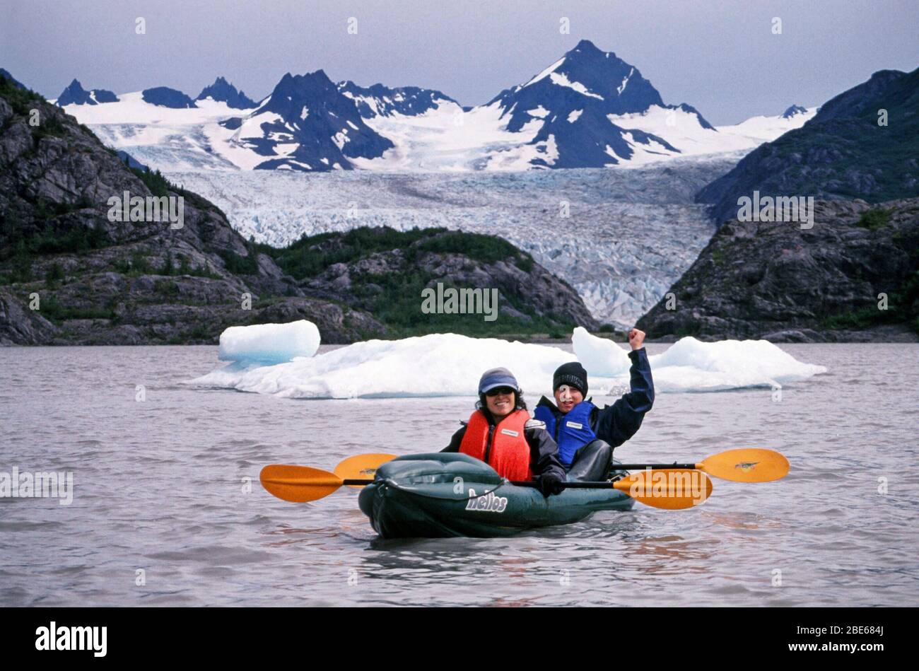 Mutter und Sohn in warmen Jacken, Hüten und Rettungswesten paddeln an einem Sommertag mit dem Kajak an Eisbergen in Halibut Cove, Homer, Alaska, USA vorbei. Stockfoto