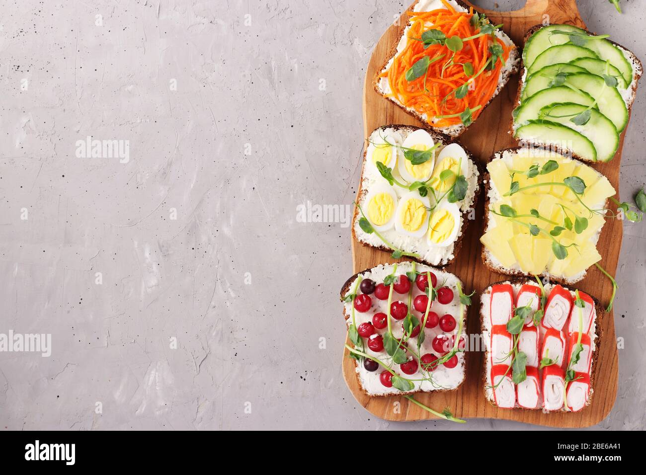 Sechs Sandwiches auf Toast mit frischen Karotten, Gurken, Ananas, rotem Currant, Krabbenstäbchen und Wachteleiern mit Erbsen Mikrogrün auf Holzbrett Stockfoto