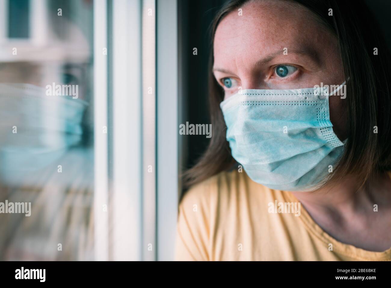 Frau in Selbstisolation während des Virenausbruchs, die durch das Fenster schaut. Besorgte weibliche Person mit schützenden chirurgischen Maske in zu Hause bleiben Konzept, sel Stockfoto