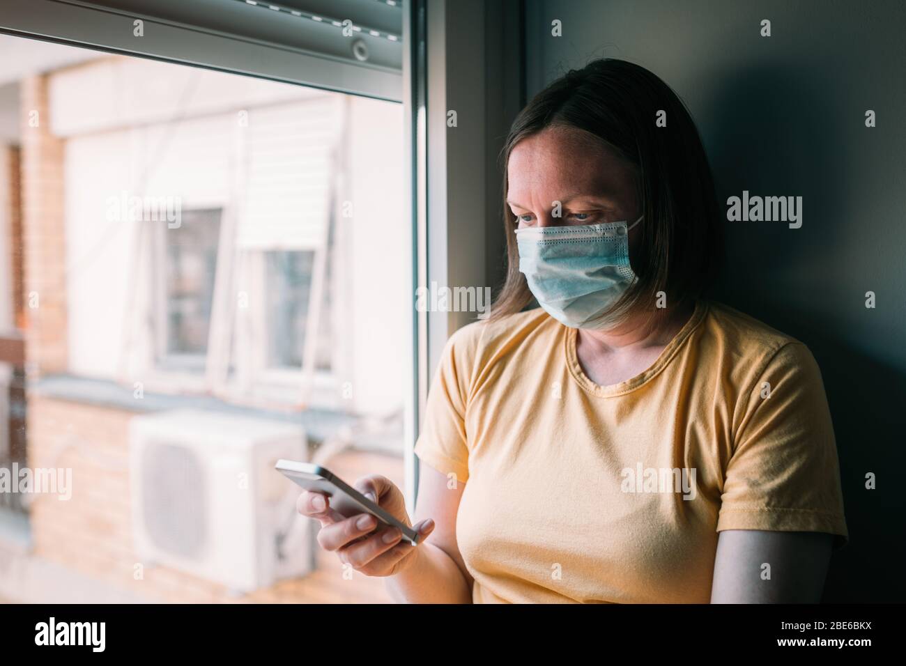 Frau in Selbstisolation während des Virus-Ausbruchs mit dem Mobiltelefon. Besorgte weibliche Person mit schützender chirurgischer Maske, hält Smartphone im Aufenthalt bei h Stockfoto
