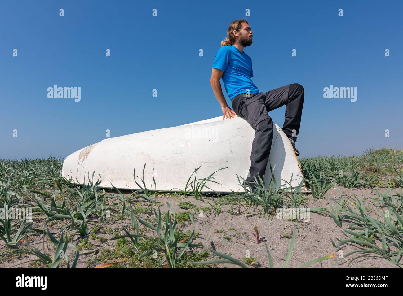 Ein Mann sitzt auf einem weißen Boot auf dem Sand. Es gibt Pflanzen im Sand. Der Himmel ist klar und blau. Stockfoto