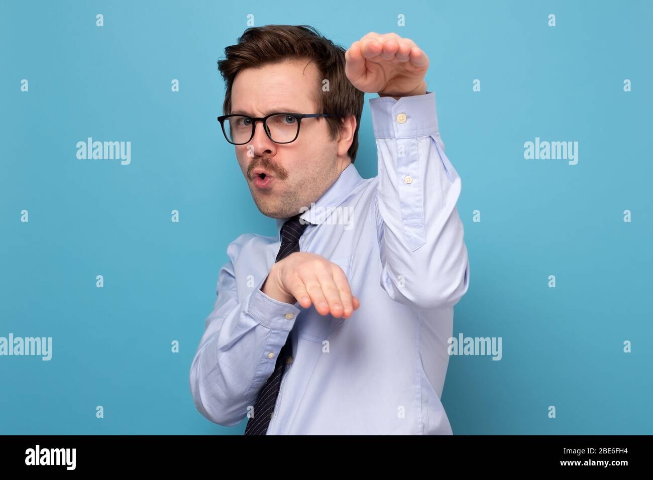 Lustig wütend schönen jungen Mann in blauem Hemd stehen in Karate Geste und bereit, anzugreifen. Innenaufnahme des Studios, isoliert auf blauem Hintergrund Stockfoto
