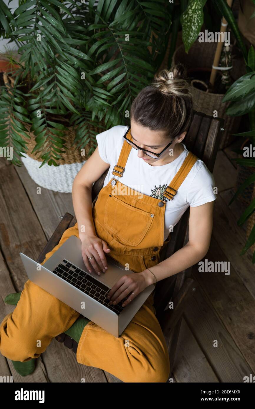 Junge Gärtnerin in Gläsern trägt Overalls, sitzt auf Holzstuhl im Gewächshaus, mit Laptop nach der Arbeit, kommuniziert im Internet mit custo Stockfoto