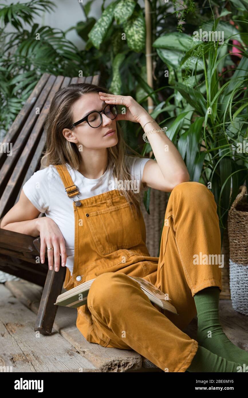 Müde Gärtnerin in Gläsern, ruhend nach der Arbeit, auf dem Holzboden sitzend, auf einem Stuhl im Haushaus gelehnt, ihre Stirn berührend, geschlossen Stockfoto
