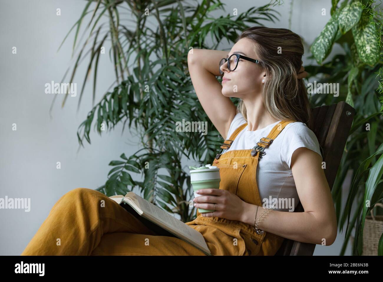 Gärtnerinnen tragen Overalls, Ruhe nach der Arbeit, sitzen auf Holzstuhl in Haus Gewächshaus, halten wiederverwendbare Kaffee / Tee Tasse, Blick auf das Fenster Stockfoto