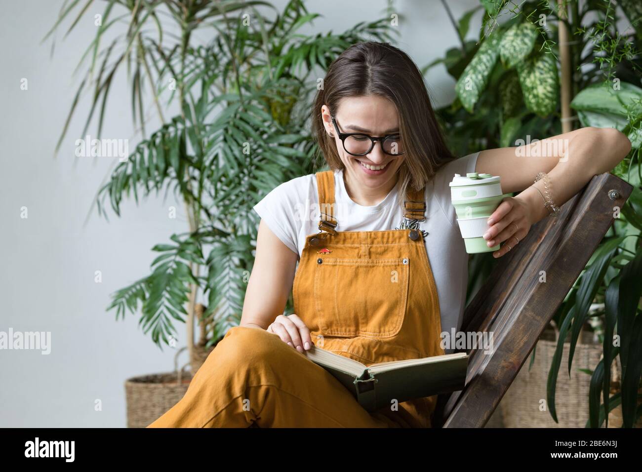Junge Gärtnerin in Gläsern tragen Overalls, Ruhe nach der Arbeit, sitzen auf Holzstuhl in Haus Gewächshaus, halten wiederverwendbare faltbare Kaffee / te Stockfoto