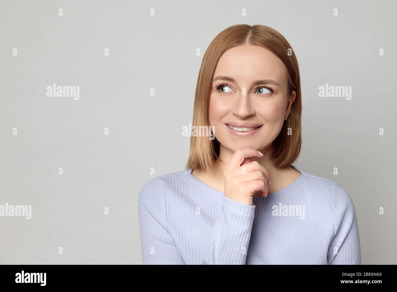 Freudige europäerin mit kurzen Haaren, in Lavendelbluse gekleidet, berührt Kinn und sieht beiseite auf leere Kopie Raum für Produkt, Denken an shoppi Stockfoto