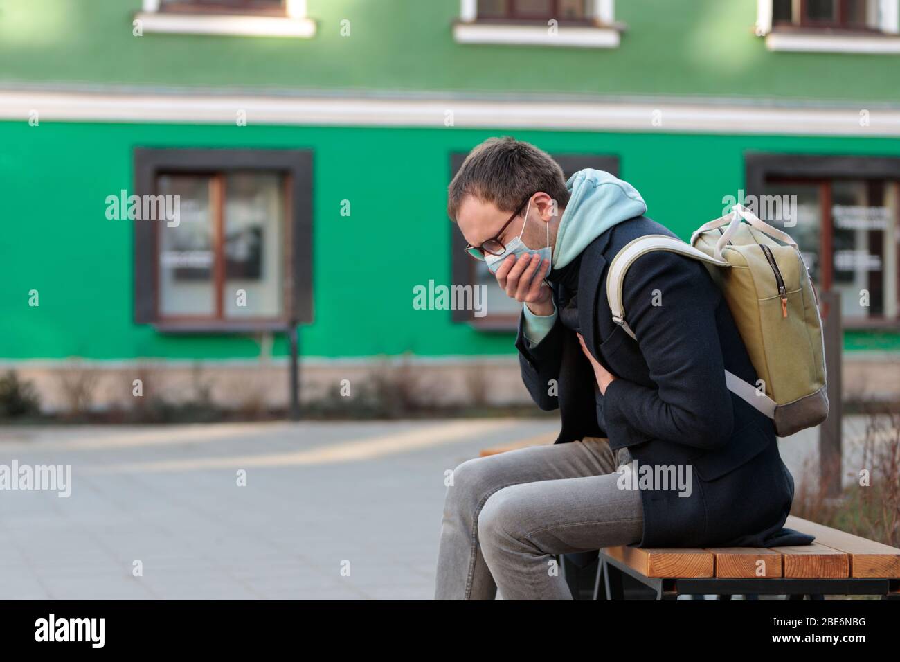 Mann in Brille krank fühlen, auf Bank sitzen, Husten, tragen Schutzmaske gegen übertragbare Infektionskrankheiten und Schutz gegen viru Stockfoto