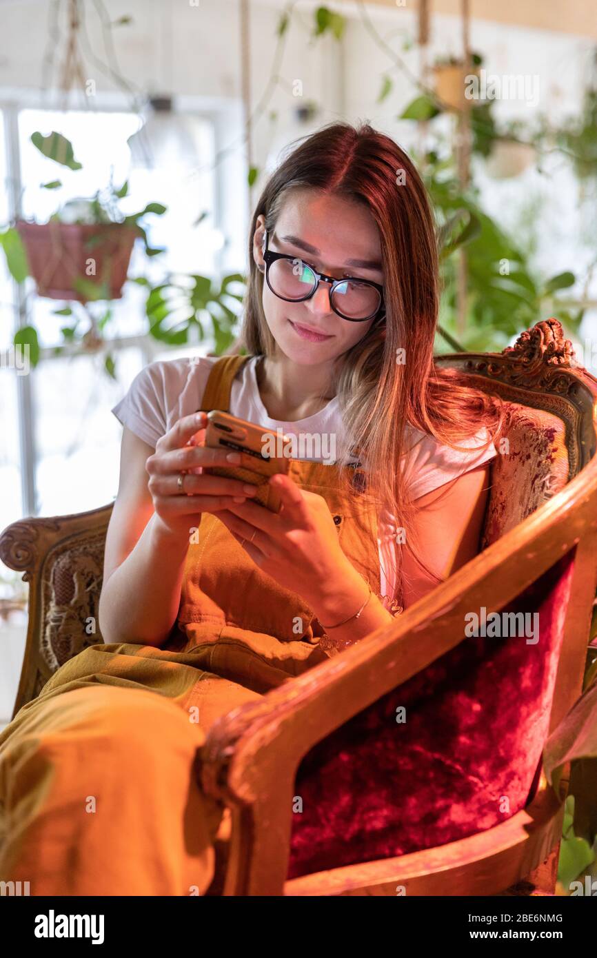 Junge Gärtnerin in Gläsern trägt Overalls, sitzt auf Vintage-Stuhl im Gewächshaus und mit Smartphone, Eingabe Nachricht, selektive Fokussierung. Werk Stockfoto