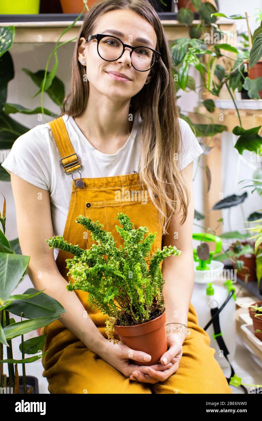 Gärtnerin in Gläsern trägt Overalls, hält einen Farn in Kunststoff-Topf, Topfpflanzen auf Hintergrund. Garten, Liebe zu Zimmerpflanzen, kostenlos Stockfoto